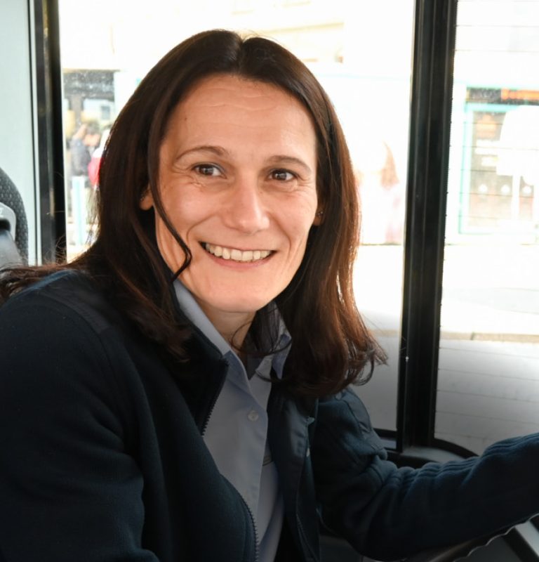 Eine Buslenkerin mit dunklen, schulterlangen Haaren sitzt am Fahrersitz eines Busses, der linke Arm liegt auf dem Lenkrad. Sie lächelt frontal in die Kamera