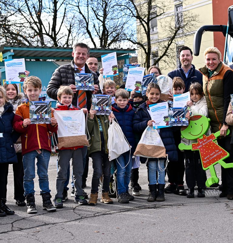 Eine Gruppe von Volksschülern sowie acht Erwachsene schauen lachend in die Kamera. Alle halten ein Buch mit dem Titel 