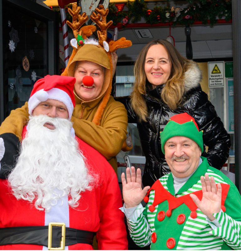 Weihnachtsmann, Rentier, Weihnachtswichtel und Projektleiterin des KMG-Weihnachtsbusses lächeln frontal in die Kamera