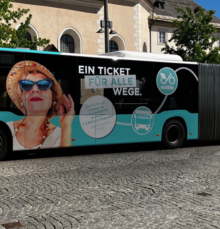 Ein KMG-Gelenksbus steht mit seiner gesamten Seitenlänge auf dem Heiligengeistplatz. Die ganze Seite des Busses ist mit einem Fullbranding beklebt. Man sieht den Kopf einer älteren Frau mit Sonnenbrille, die mit ihrer linken Hand einen Strohhut auf dem Kopf hält. Daneben ist ein weißer Kreis mit der Info zu den inkludierten Angeboten der KMG-Jahreskarte PLUS sowie der Schriftzug 