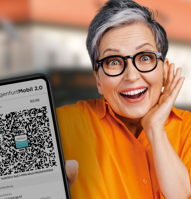 Frau lächelt in die Kamera und hält ein Handy entgegen, auf dem der QR-Code für die KlagenfurtMobil 2.0-App ersichtlich ist. Im Hintergrund ist verschwommen das Gebäude des Landhaushofes am Heiligengeistplatz in Klagenfurt zu sehen