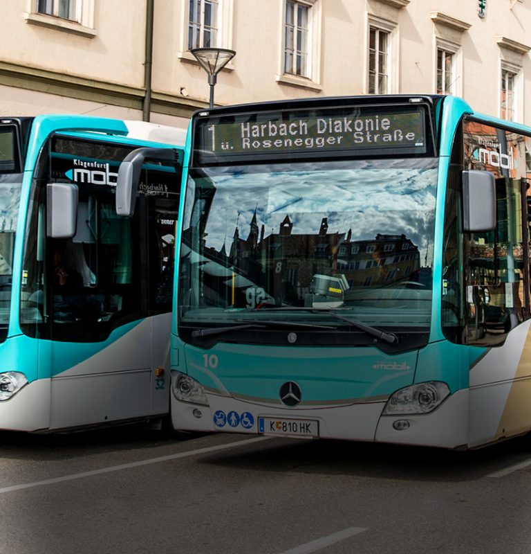Zwei Busse fahren nebeneinander, zu sehen sind deren Fronten sowie leicht schräg die Fahrerseite des rechten Busses. Links dahinter sieht man eine Häuserfront.