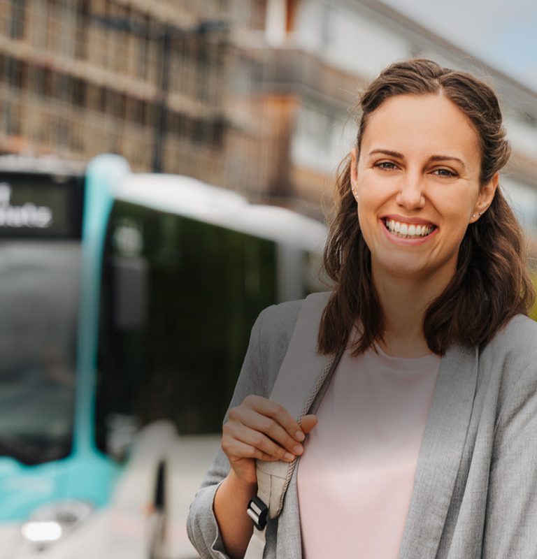 Junge Frau mit dunklen, schulterlangen Haaren lächelt in die Kamera. Links hinter ihr sieht man einen Bus der in Richtung Kamera fährt, dahinter befindet sich verschwommen ein im Bau befindliches Gebäude mit Baugerüst davor.