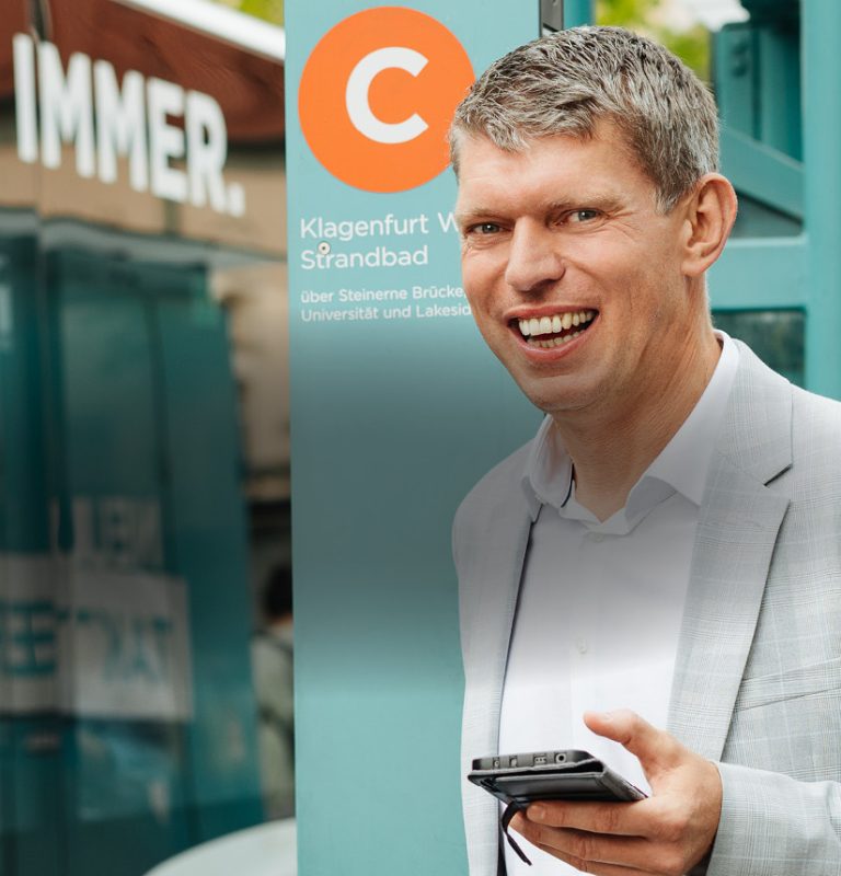 Mann im mittleren Alter steht bei einer Bushaltestelle, lächelt in die Kamera und hält ein Smartphone in seiner linken Hand. Dahinter ist die Haltestellenstele mit einem runden, orangen 