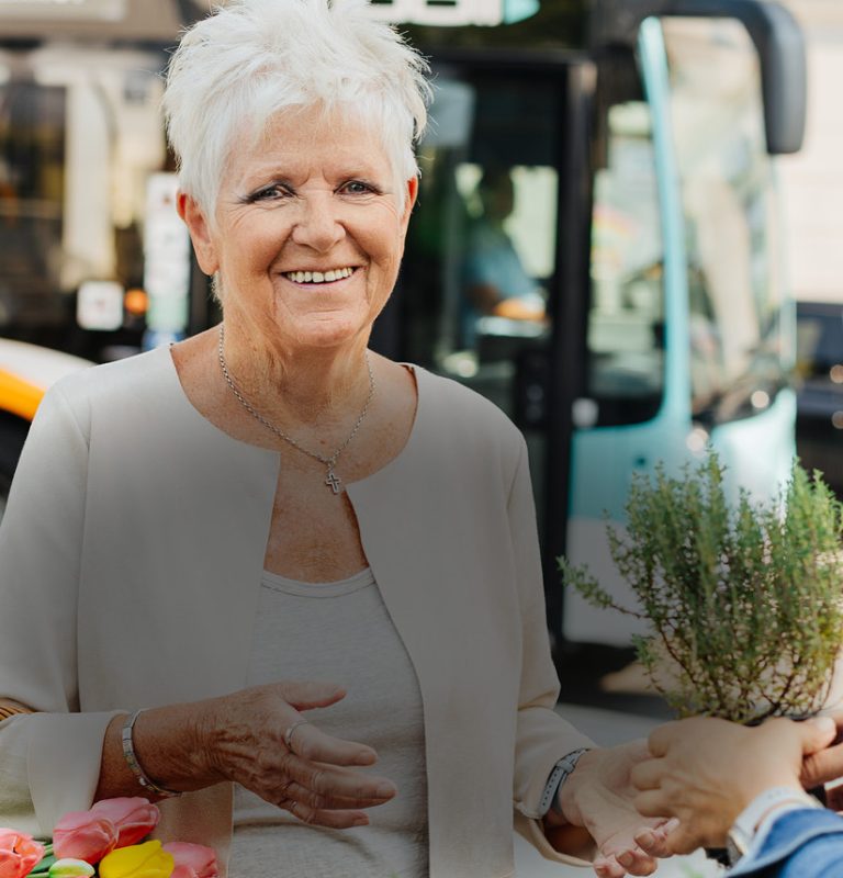 Im Hintergrund sieht man einen KMG-Bus von der Seite. Im Vordergrund sieht man eine ältere Frau mit kurzen, weißen, Haaren. Auf ihrem abgewinkelten, rechten Arm befindet sich ein Korb mit einem bunten Tulpenstrauß. Links danebe sieht man einen Strauß mit grünen Kräutern. Sie lächelt in die Kamera. Von rechts sieht man eine Person mit längeren brünetten Haaren, die ihr einen Topf mit Kräutern entgegenstreckt.