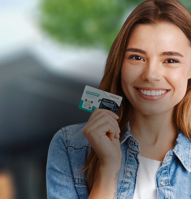 Junge Frau lacht in die Kamera und hält dabei eine KMG-Prepaid Card in der rechten Hand. Im Hintergrund sieht man verschwommen das Dach einer Bushaltestelle sowie die Blätter von Bäumen.