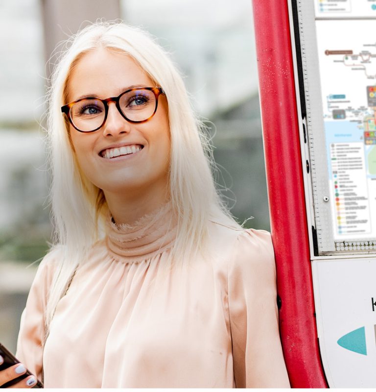Blonde junge Frau mit dunkler Brille hält Handy in der Hand und schaut seitlich an der Kamera vorbei auf den Fahrplan an der Haltestellen-Stele.