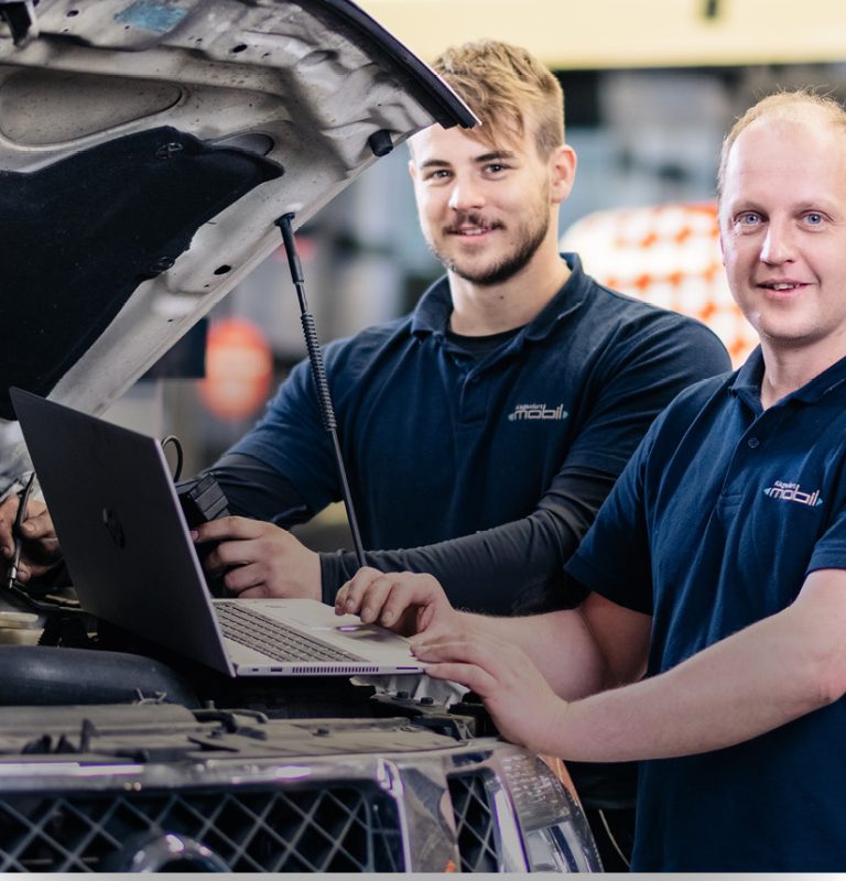 Zwei junge Männer stehen hinter der offenen Motorhaube eines Fahrzeuges. Der vordere der beiden bedient einen Laptop, der auf der Motorhaube aufliegt. Beide lächeln in die Kamera.