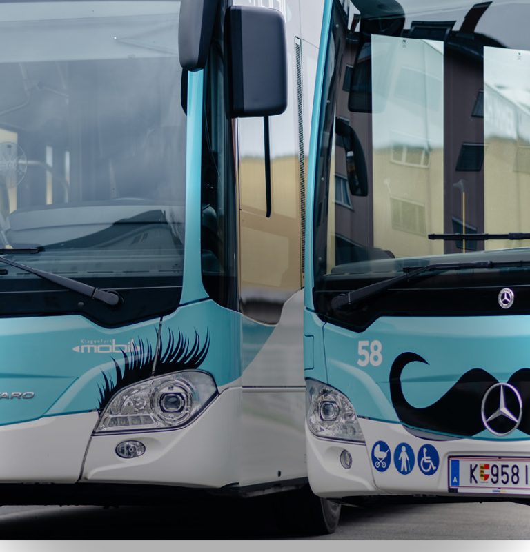 Man sieht die Fronten von zwei nebeneinanderstehenden KMG-Bussen. Der linke hat über den Scheinwerfern eine Beklebung in Form von Wimpern, der rechte in der Mitte eine Beklebung in Form eines Schnauzbartes.