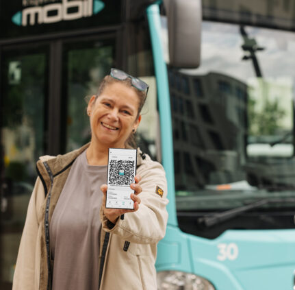 Eine Frau steht lächelnd vor einem KMG-Bus und hält mit ausgestrecktem Arm ihr Smartphone mit einem Screenshot der KlagenfurtMobil 2.0-App in die Kamera