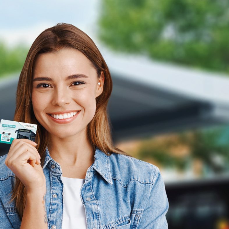 Junge Frau lacht in die Kamera und hält dabei eine KMG-Prepaid Card in der rechten Hand. Im Hintergrund sieht man verschwommen das Dach einer Bushaltestelle sowie die Blätter von Bäumen.