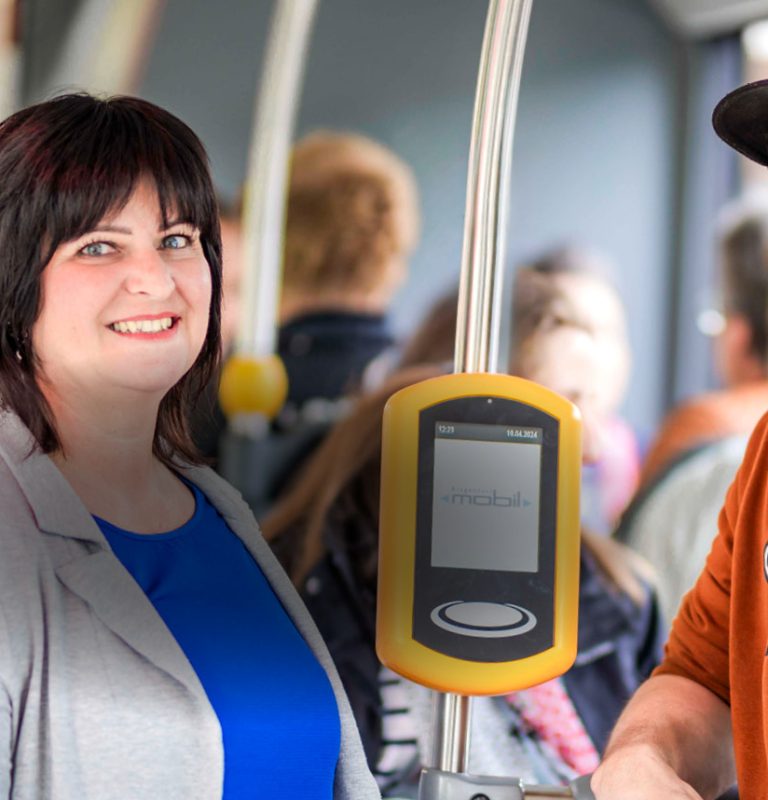 Eine Frau und ein Mann stehen nebeneinander im KMG-Bus und lächeln in die Kamera. Zwischen den beiden befindet sich eine Haltestange, auf der ein Entwerter montiert ist.