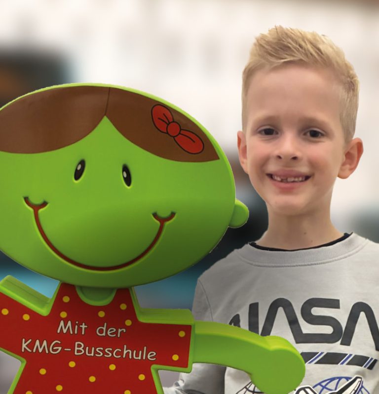 Ein blonder Junge mit grauem Nasa-T-Shirt lächelt in die Kamera und zeigt mit seiner linken Hand das Zeichen für Daumen hoch. Links neben ihm ist im Vordergrund die grüne Kunststoff-Figur von Hanna Haltefest, mit seitlilch weggestreckten Armen zu sehen.