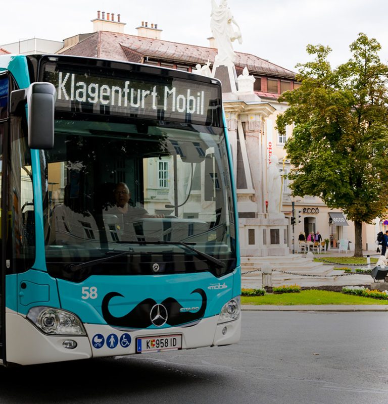 Türkiser KMG-Bus mit Frontbeklebung in Form eines Schnauzbartes fährt von links kommend in den Kreisverkehr am Heuplatz in Klagenfurt ein.