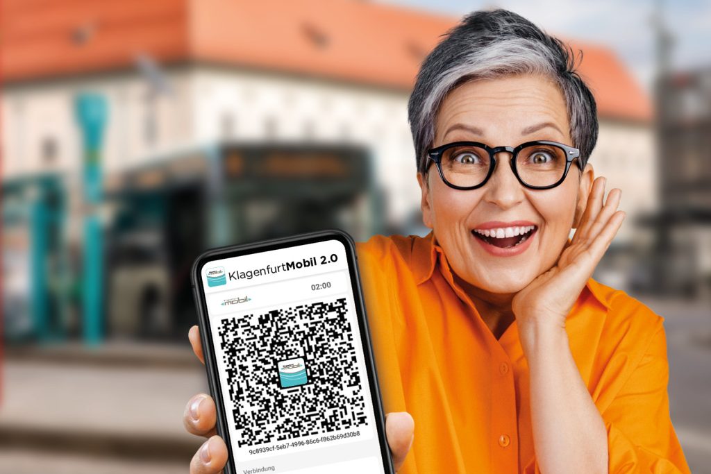Frau lächelt in die Kamera und hält ein Handy entgegen, auf dem der QR-Code für die KlagenfurtMobil 2.0-App ersichtlich ist. Im Hintergrund ist verschwommen das Gebäude des Landhaushofes am Heiligengeistplatz in Klagenfurt zu sehen